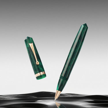 Una penna verde traslucida con dettagli dorati, raffigurata senza cappuccio e sospesa sopra una superficie scura e riflettente, su uno sfondo grigio sfumato.