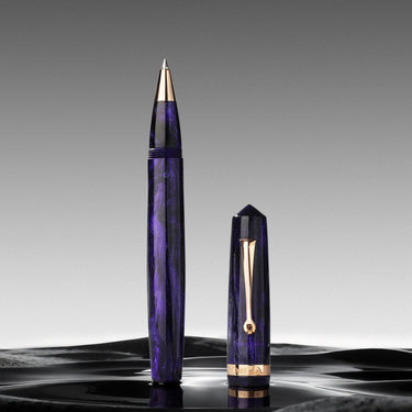 Una penna roller di lusso con il suo cappuccio su una superficie scura e riflettente, davanti a uno sfondo grigio sfumato. La penna presenta un corpo in resina marmorizzata viola scuro con dettagli placcati in oro sulla punta, sulla clip e alla base.