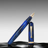Una penna roller di lusso in blu marmorizzato con dettagli dorati, raffigurata con il cappuccio rimosso e posto in verticale accanto al corpo, su una superficie nera lucida e uno sfondo grigio sfumato.